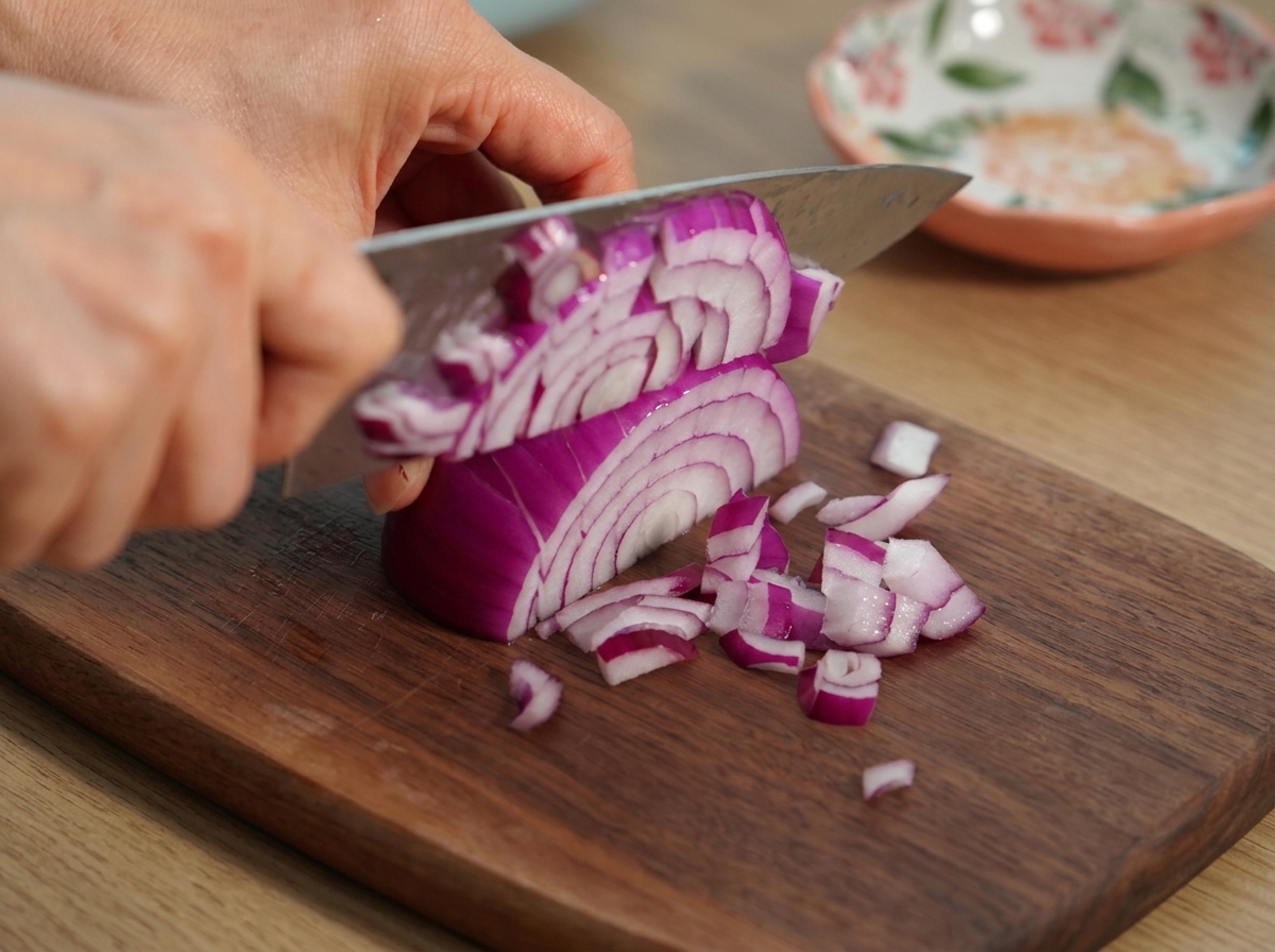 Hands using a chefs knife to chop a red onion into fine pieces on a wooden cutting board.