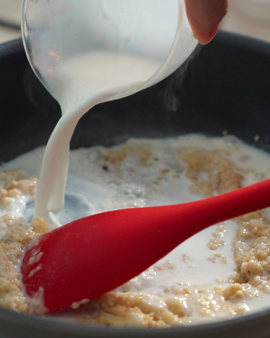 Milk being poured from a glass measuring cup into a dark pan containing a thick butter and flour paste, being stirred with a red silicone spatula.