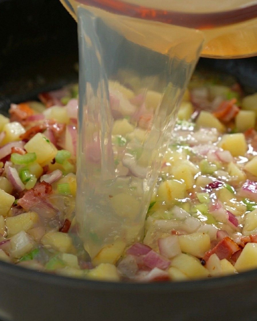 Fresh black pepper being ground over a pan of simmering clam broth and cooked vegetables.