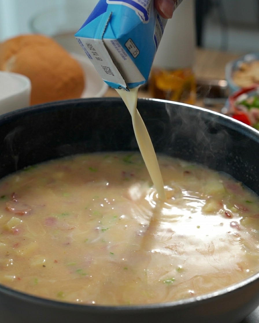 Heavy cream being poured from a carton into a pan of simmering clam chowder base.