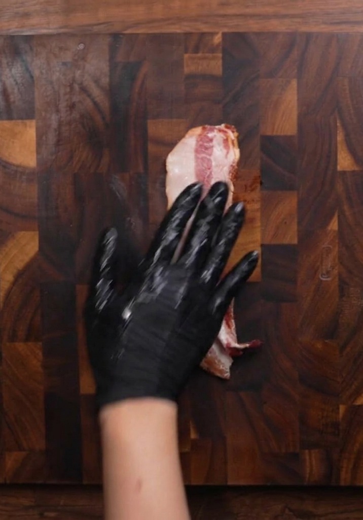A gloved hand resting on a piece of raw bacon on a dark wooden cutting board, preparing to slice it.