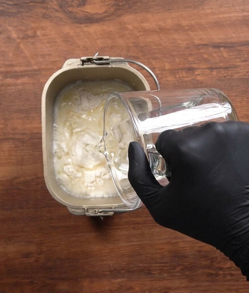 Water being poured from a clear glass measuring cup into a bread maker pan filled with flour and other ingredients.