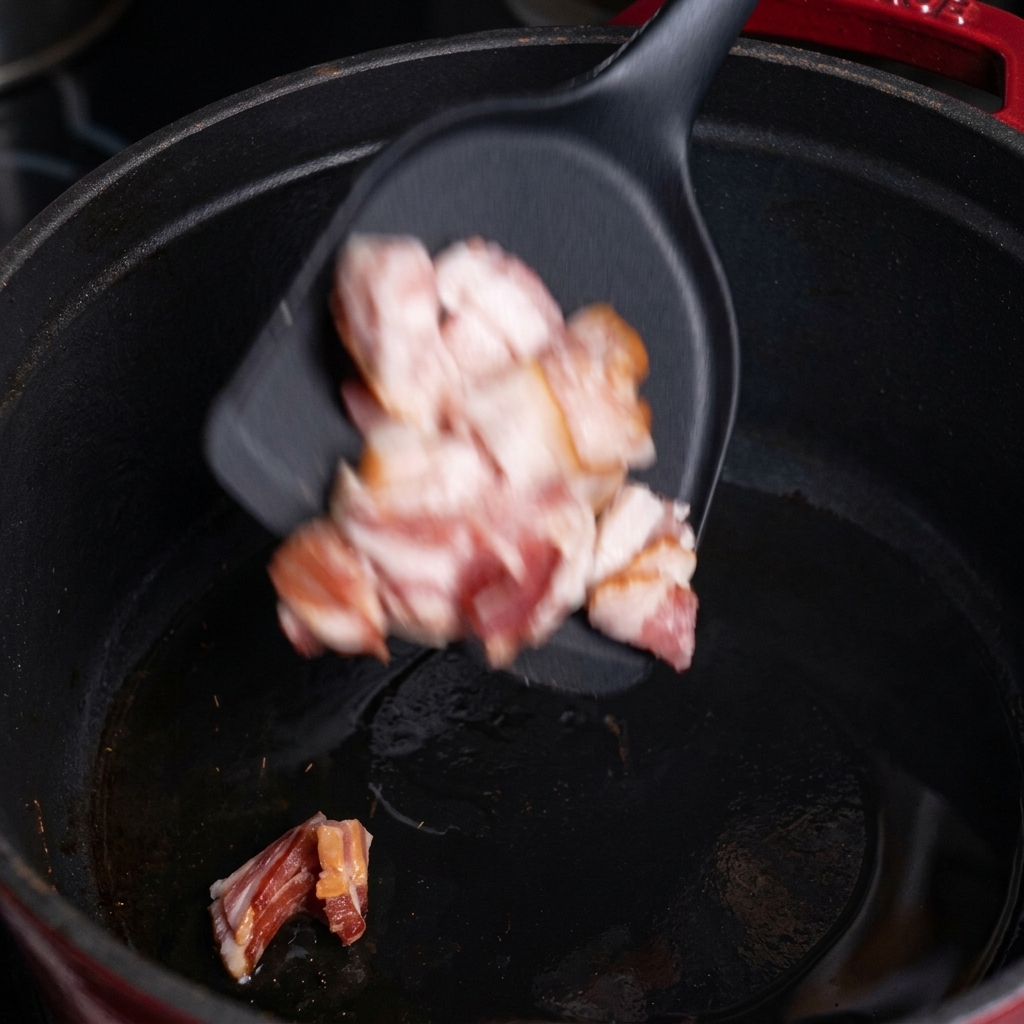 Chopped raw bacon pieces falling from a black spatula into a hot black cast iron pot.