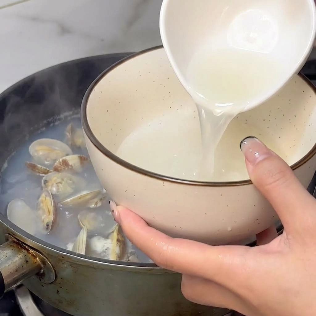 A hand using a small white bowl to scoop steaming clam broth out of a pan of cooked clams.