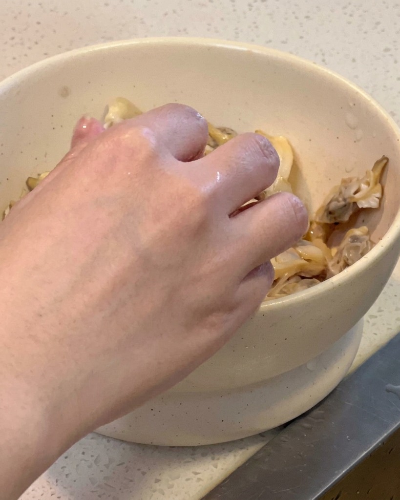 A persons hand removing cooked clam meat from its shell into a white bowl.