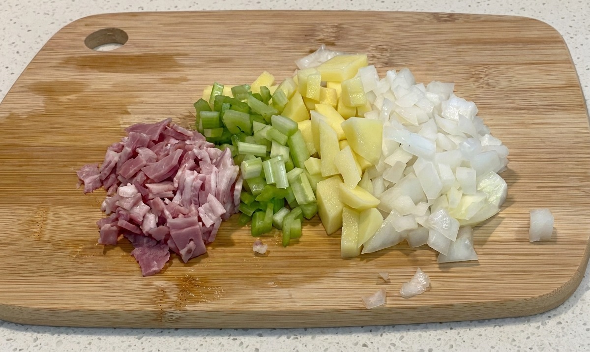 Piles of diced bacon, green celery, yellow potatoes, and white onions arranged neatly on a wooden cutting board.