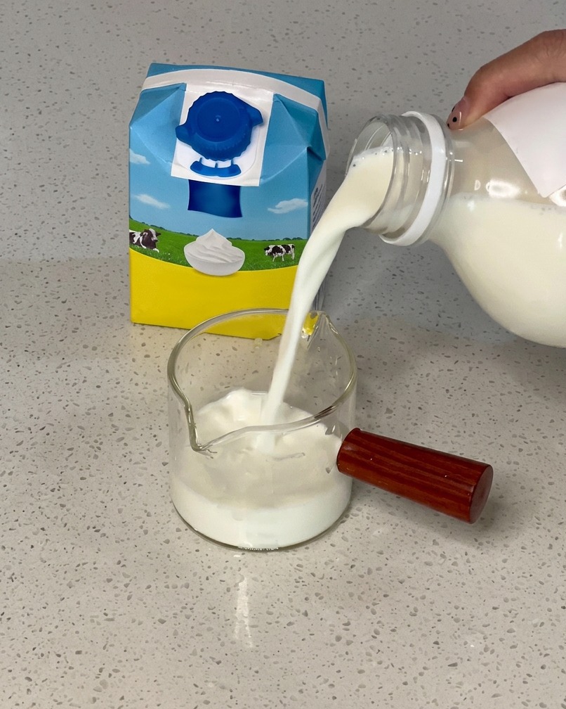 Hand pouring milk from a blue and yellow carton into a small glass pitcher filled with cream.