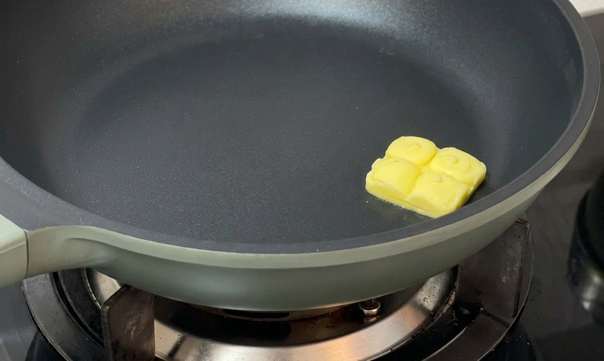 A single square pat of butter melting in the center of a dark non-stick skillet.