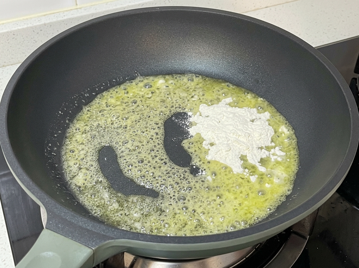 A grey pan on a stovetop containing bubbling melted yellow butter with a pile of white flour being stirred in.