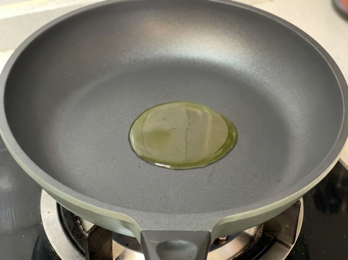 A small pool of golden olive oil in the center of a clean, dark grey frying pan on a gas stove.