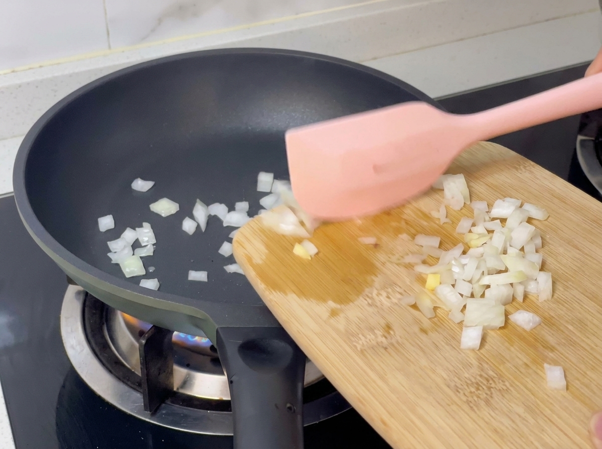 Diced white onions being scraped from a wooden cutting board into a hot pan with a pink spatula.