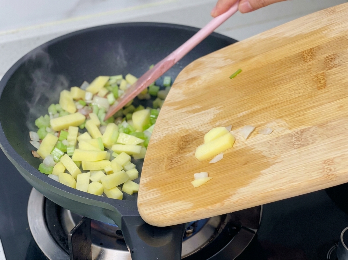 Diced potatoes being added from a wooden board into a pan already containing sautéed celery, onions, and bacon.
