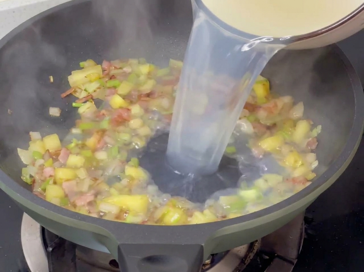 Pouring light-colored clam broth into a hot pan with sautéed diced potatoes, celery, onions, and bacon.