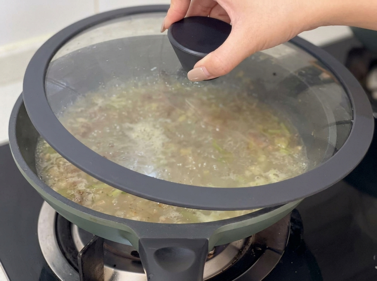 A hand placing a glass lid onto a pan of simmering clam chowder soup base.