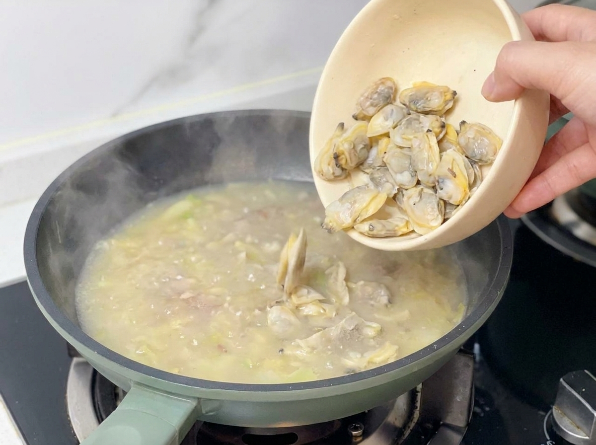 A hand pouring cooked, shelled clam meat from a beige bowl into a pan of simmering chowder base.