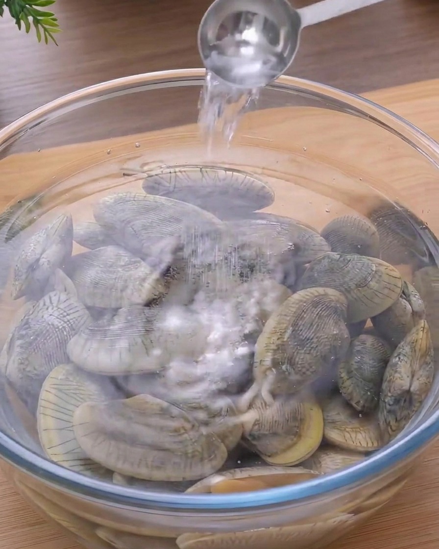 A metal spoon pouring salt into a clear glass bowl filled with water and fresh clams.