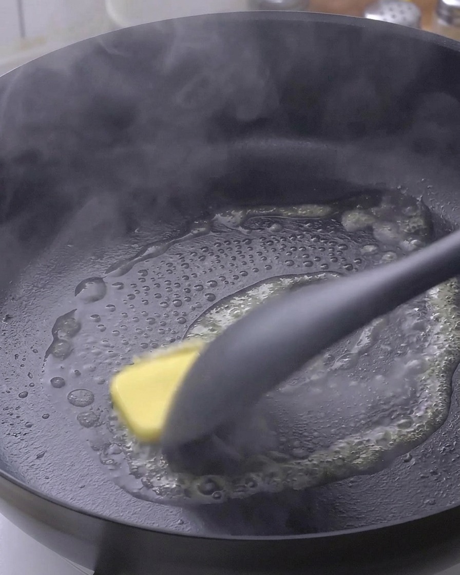 A square of yellow butter melting in a hot black frying pan, being moved around by a black spatula.