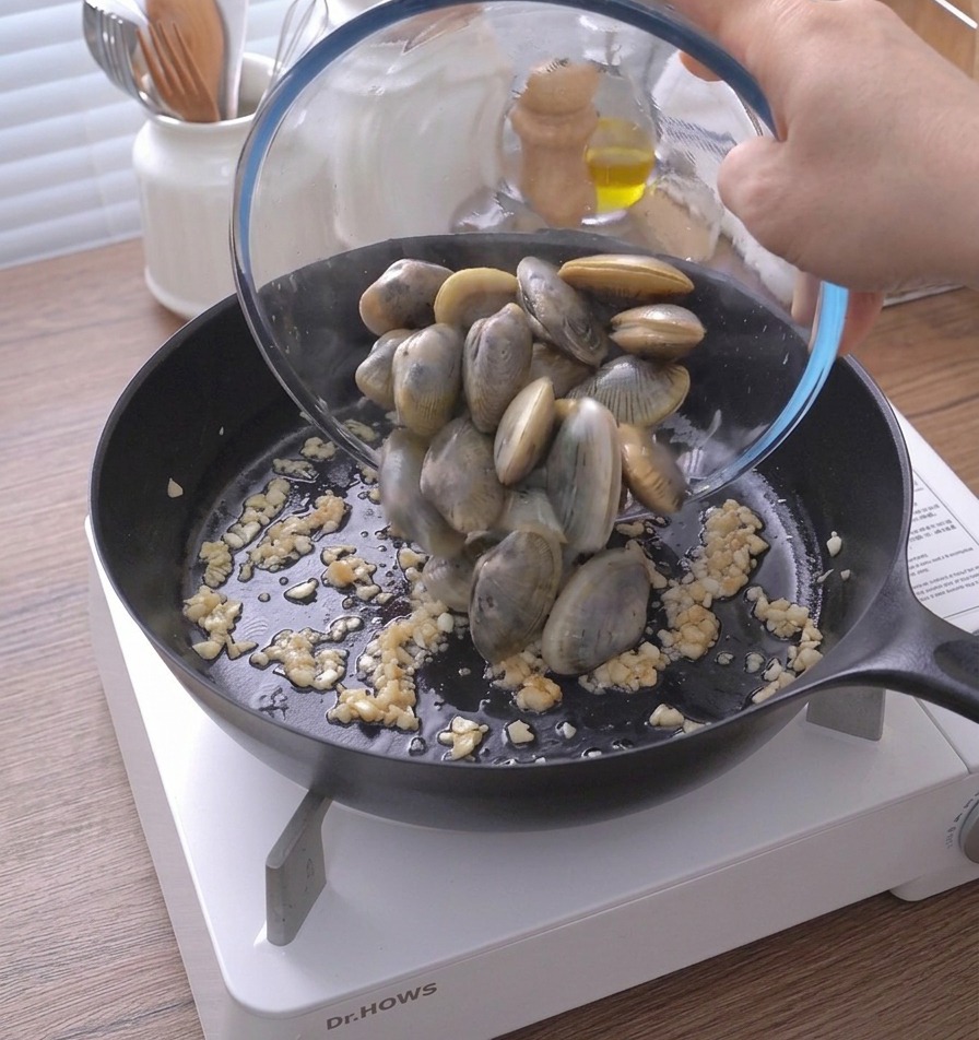Pouring whole clams from a glass bowl into a hot pan containing sautéed minced garlic and butter.