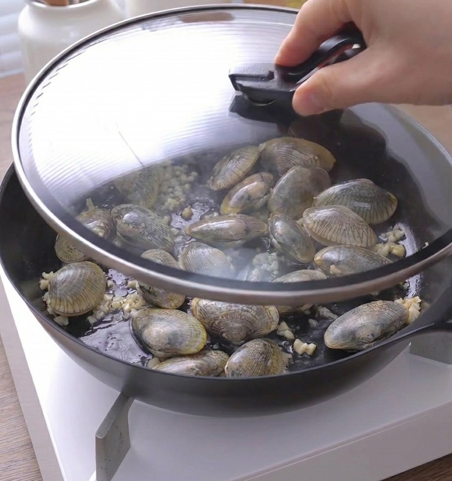 A hand placing a glass lid onto a pan full of clams and garlic.
