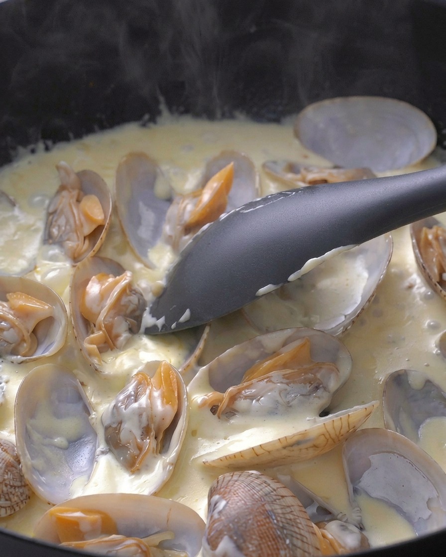 A black spatula gently stirring open clams in a bubbling white cream sauce inside a pan.