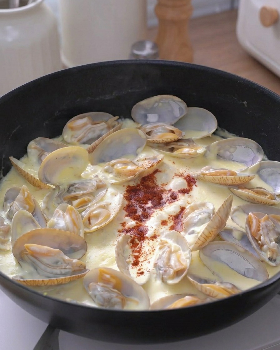 A dash of red spice powder sprinkled in the center of a pan of creamy simmering clams.