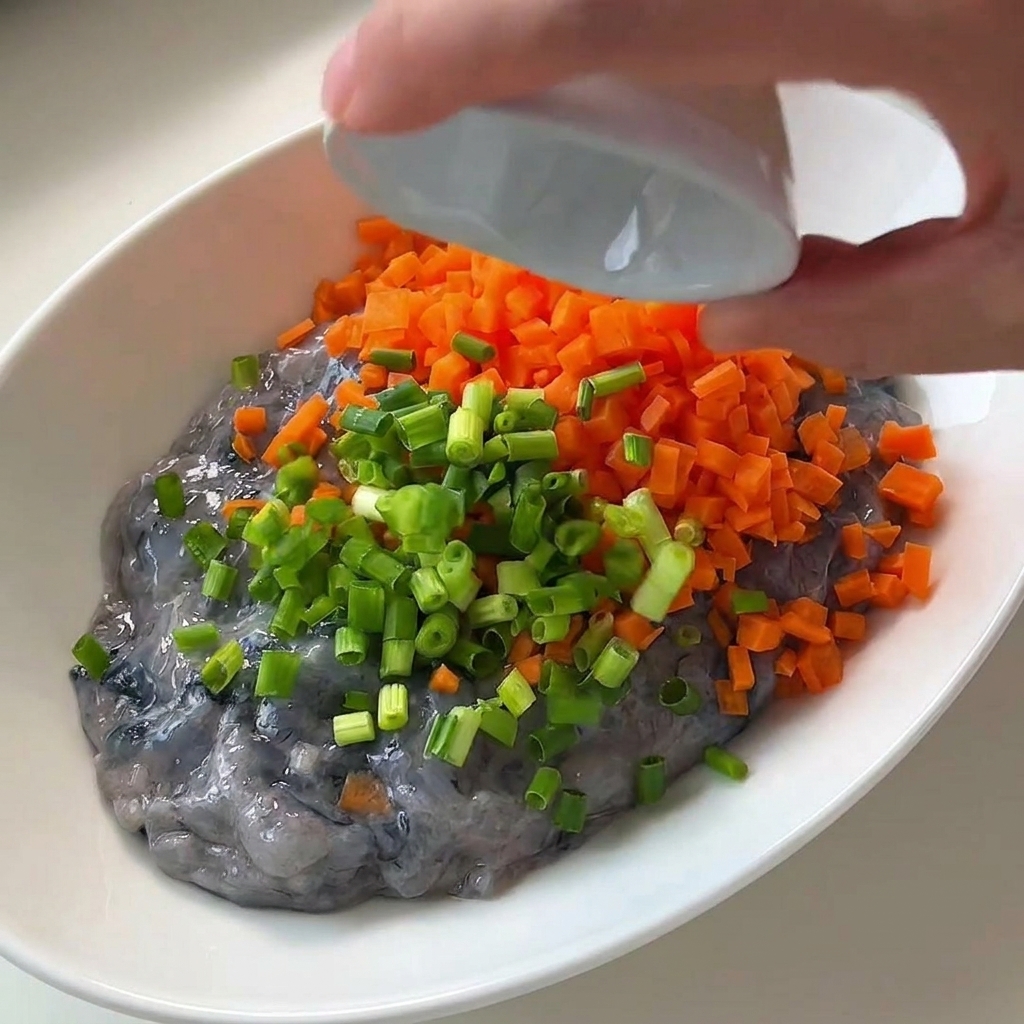 Chopped green onions being poured onto the shrimp paste and diced carrots in a white bowl.