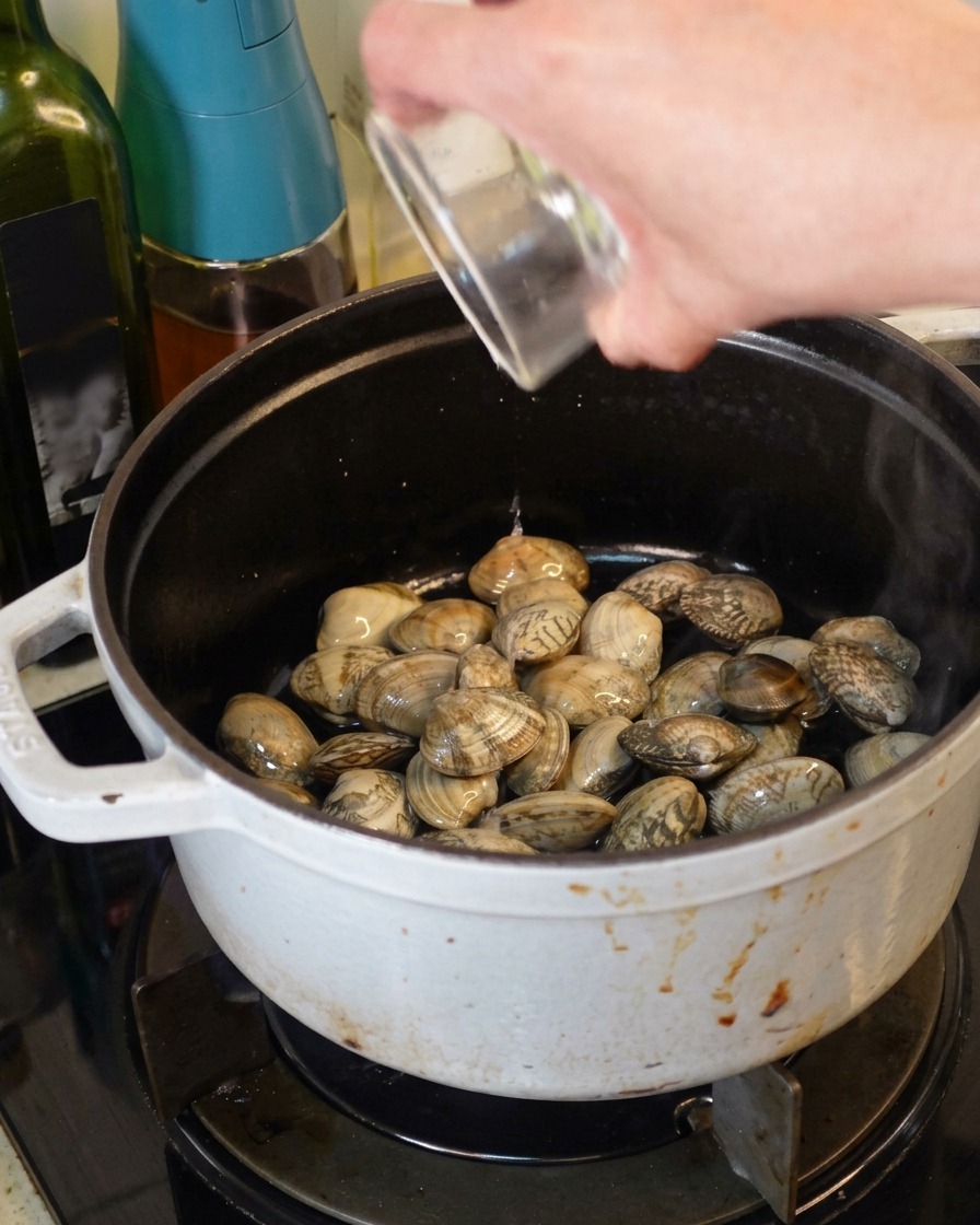 Pouring clear white wine from a small glass container into a white cast iron pot filled with fresh clams.