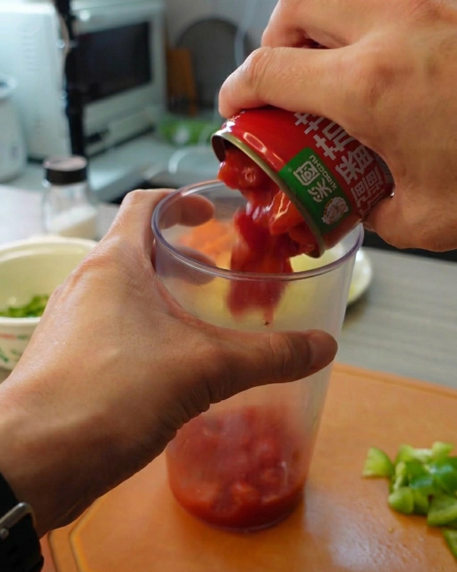 Hands pouring chopped canned tomatoes from a tin into a tall clear plastic blending cup.