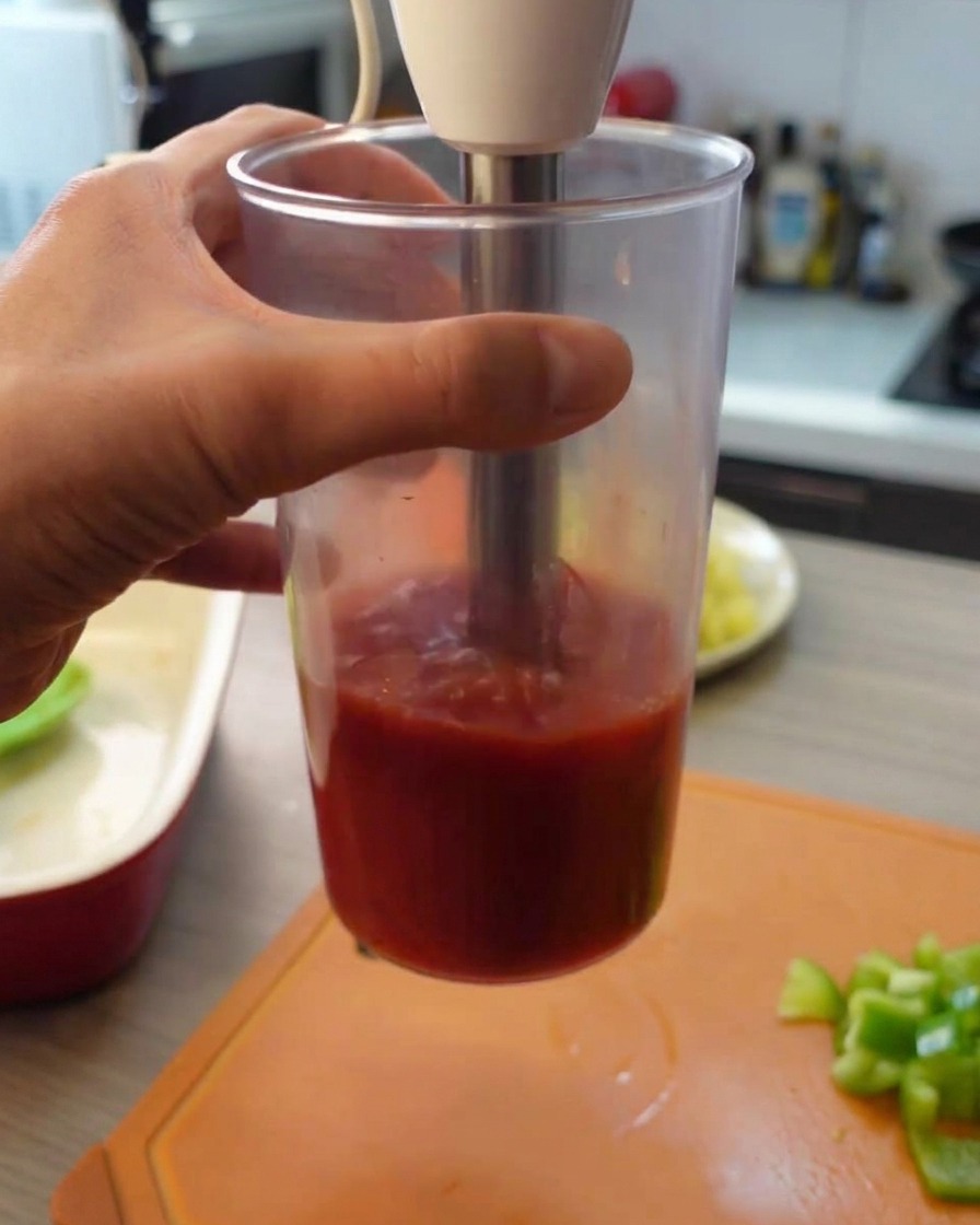 Using an immersion hand blender to purée canned tomatoes inside a tall clear plastic cup.