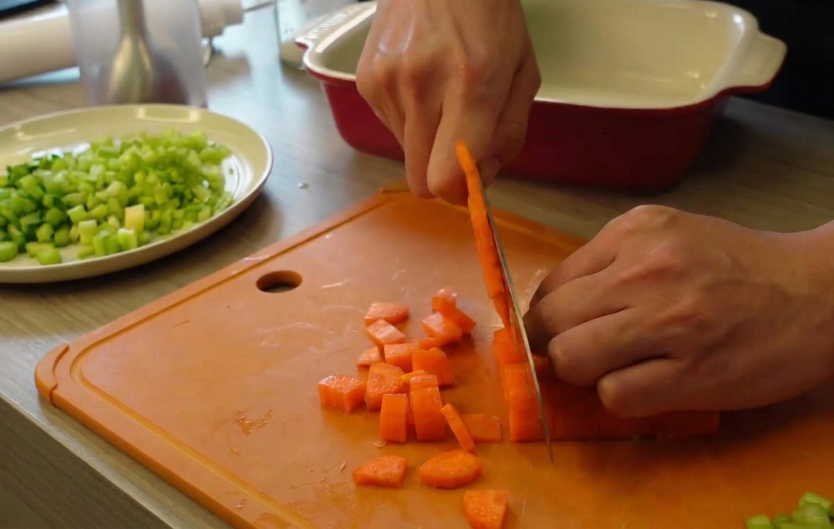 Hands using a kitchen knife to dice bright orange carrots into small cubes on an orange cutting board, with a plate of chopped celery in the background.