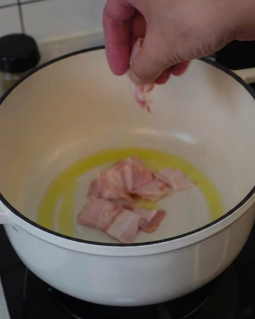 A hand placing pieces of raw bacon into a white pot with oil to begin frying.