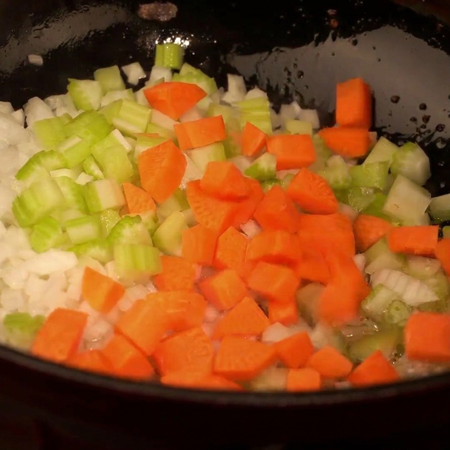 A top-down view of a black pan filled with chopped white onions, green celery, and bright orange carrot cubes being sautéed.