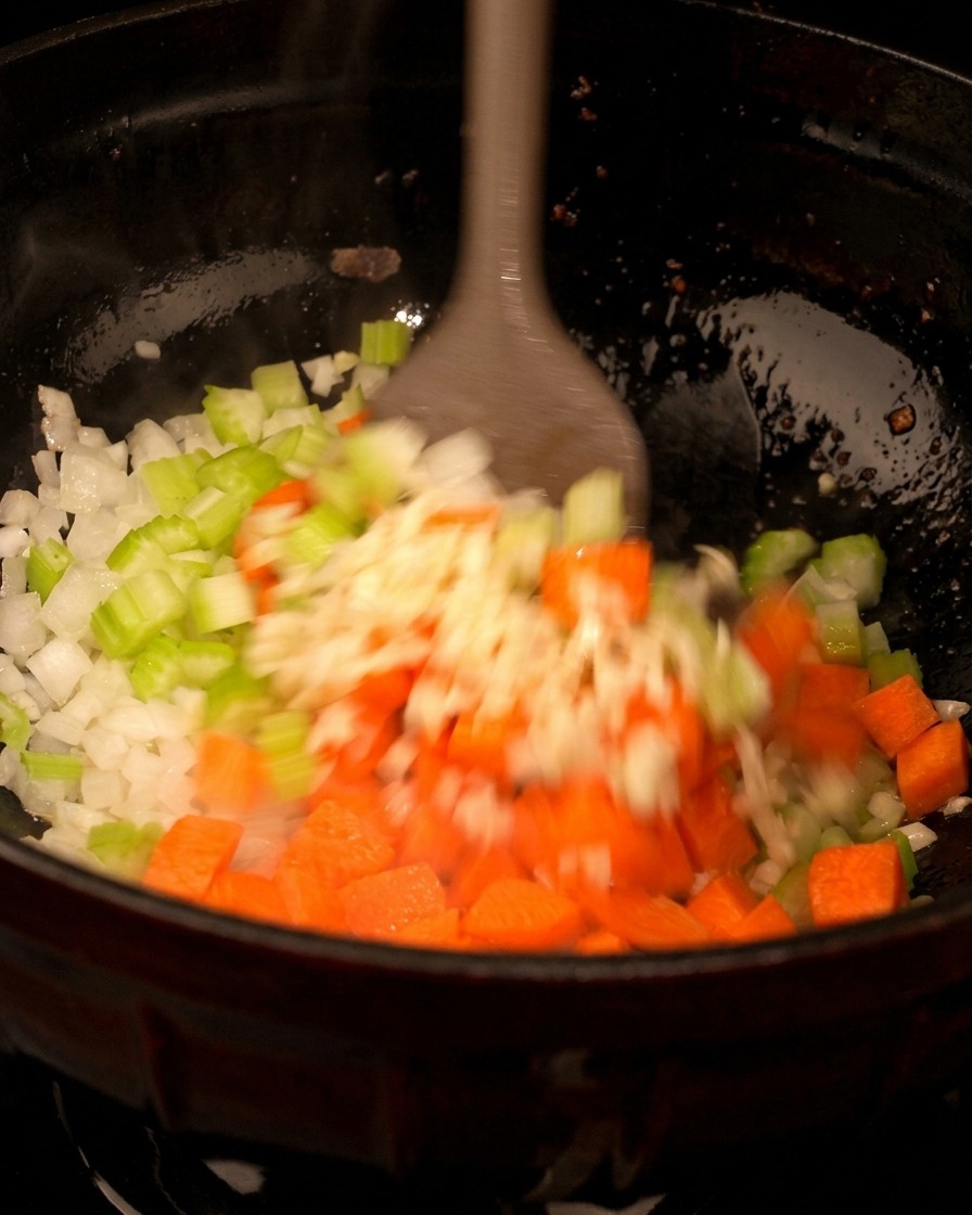 Diced carrots being tossed into a black pan containing sautéed onions and celery.