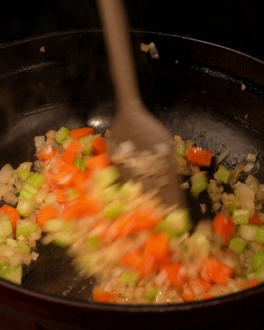 A wooden spatula actively stirring a colorful mix of diced carrots, celery, and onions in a black pan.
