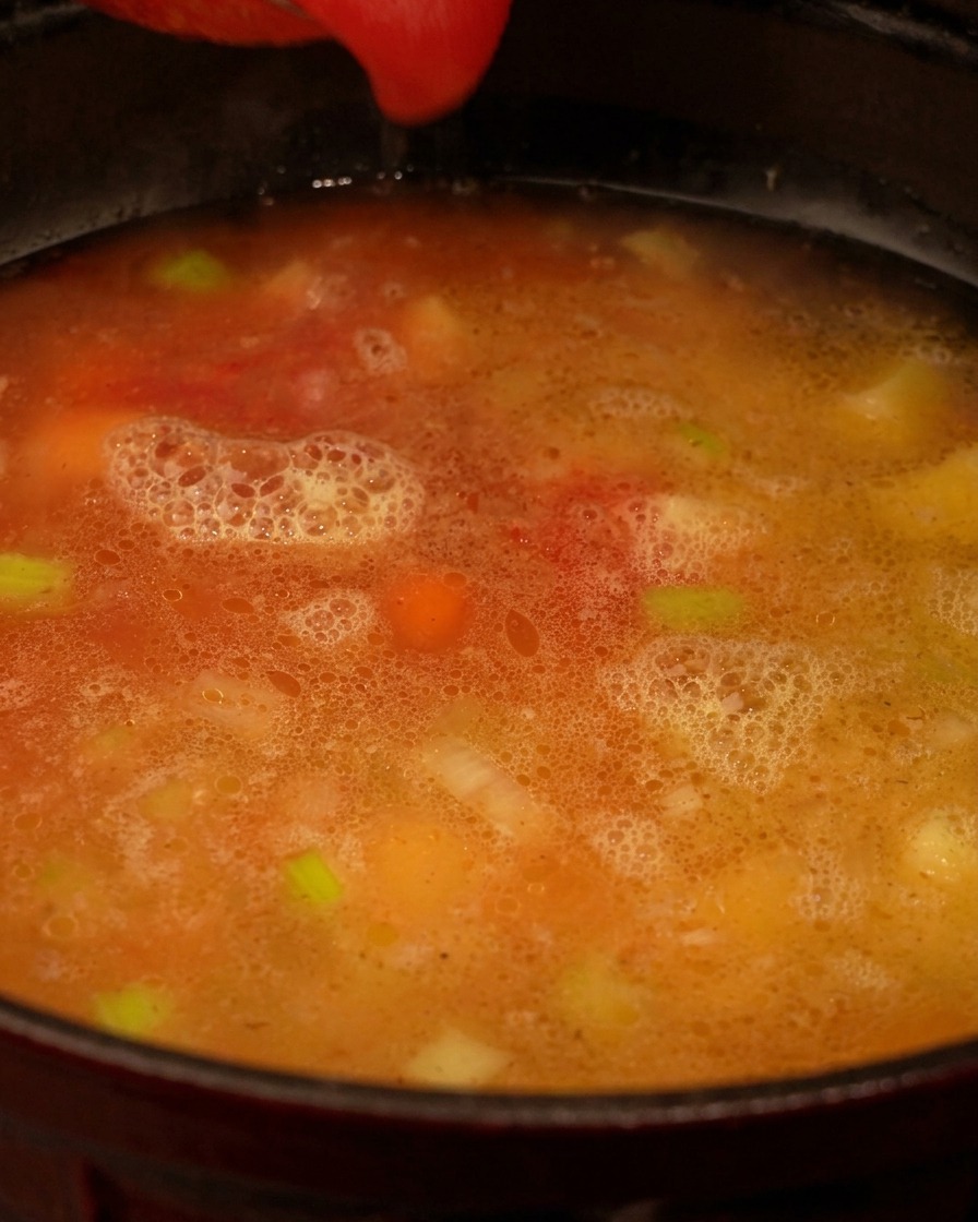 Red tomato puree being poured into a pot containing a light-colored broth with visible pieces of celery and carrots.