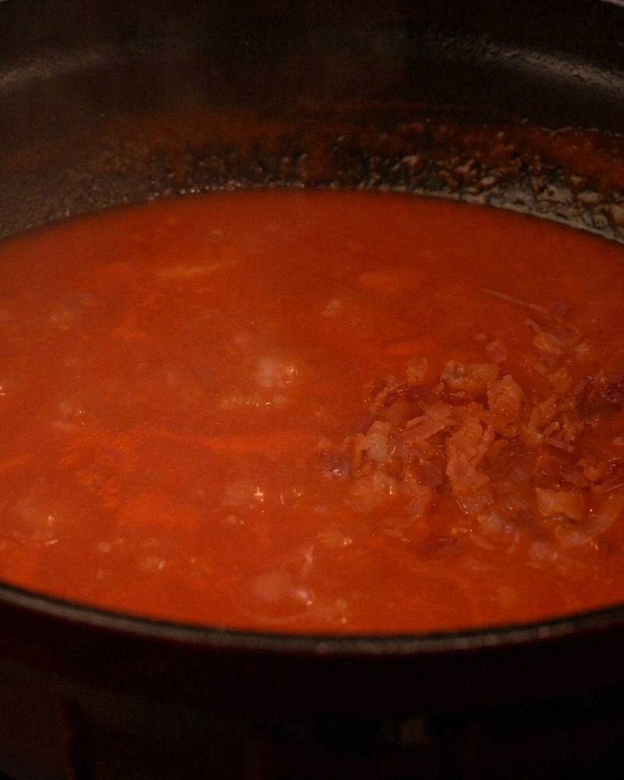 Crispy cooked bacon bits being added to a bubbling pot of rich red tomato soup.