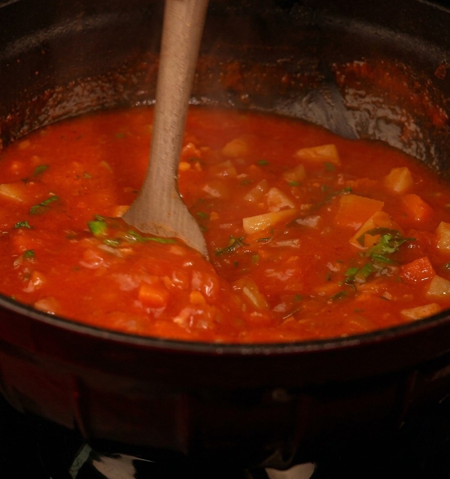 A wooden spoon stirring a thick red tomato based soup filled with diced vegetables and flecks of fresh green parsley
