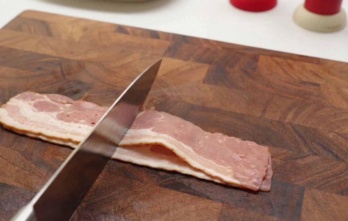 Hands using a sharp knife to slice raw strips of bacon into small pieces on a wooden cutting board.