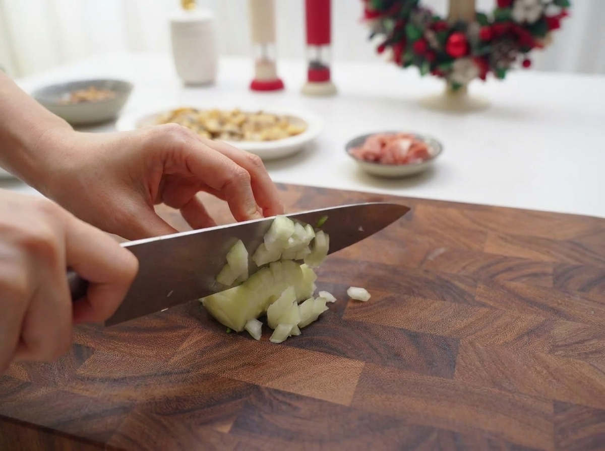 Hands using a chefs knife to dice a white onion into fine pieces on a wooden cutting board.