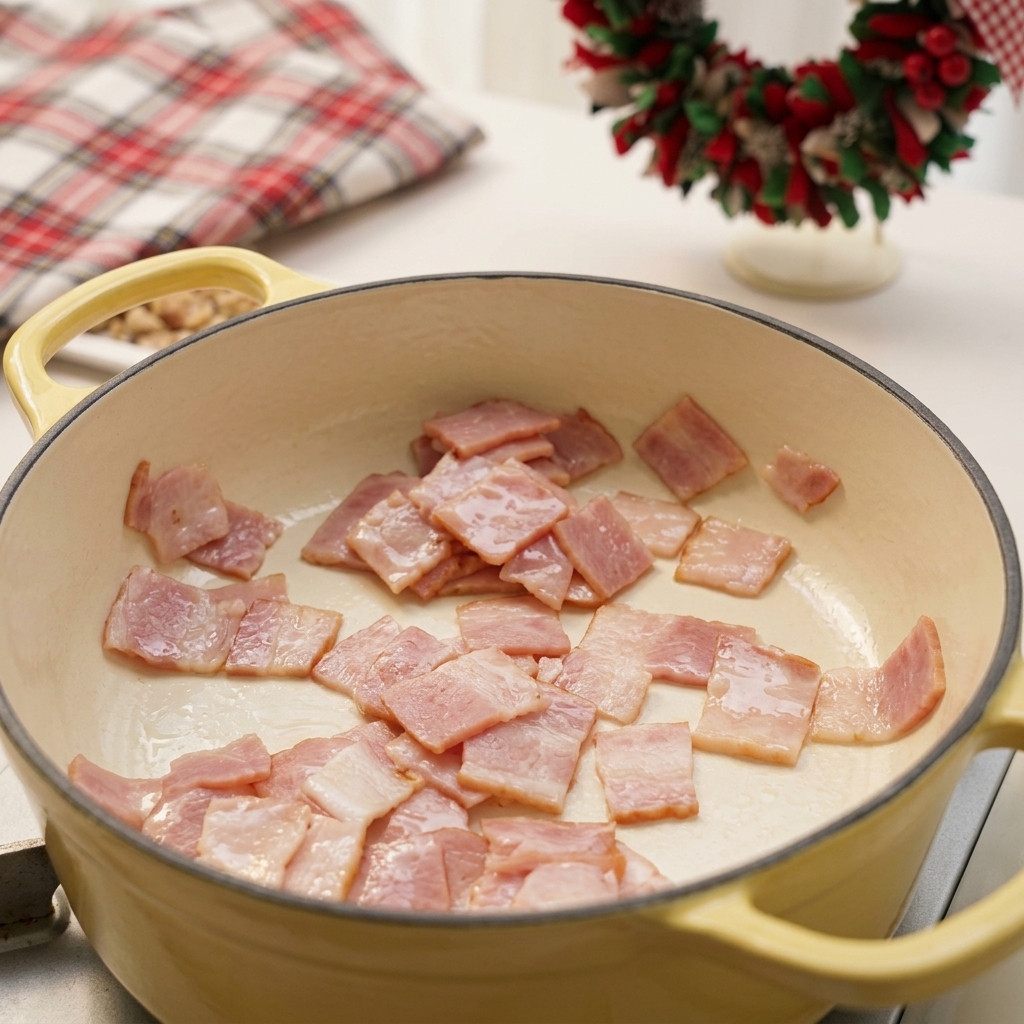 Chopped squares of bacon frying and rendering fat in a pale yellow enamel pot.