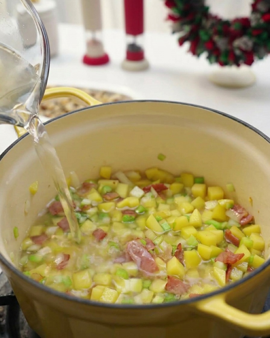 Clear liquid being poured from a glass pitcher into a yellow pot containing diced vegetables and bacon.