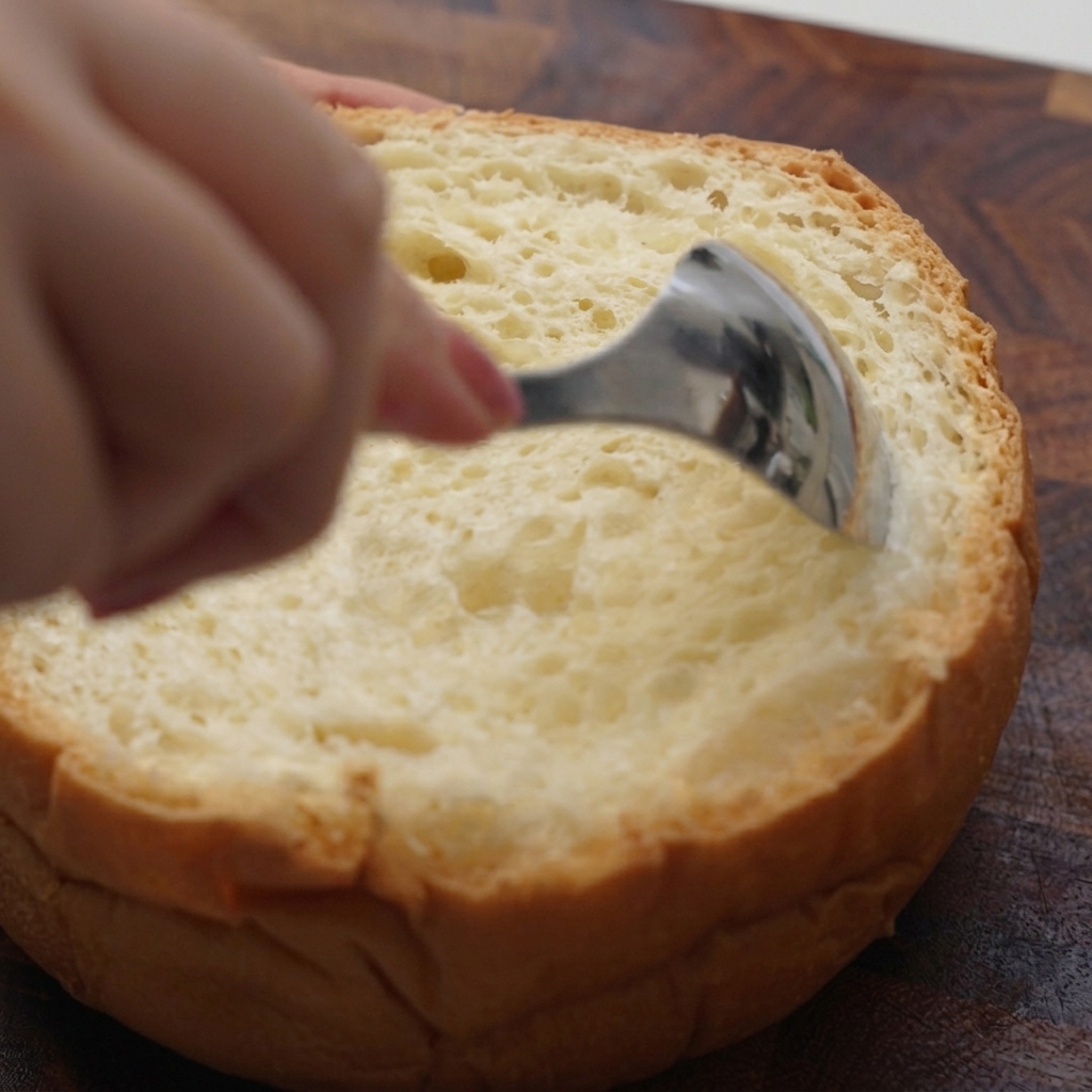 A hand using a metal spoon to scoop out the soft interior of a round loaf of bread.