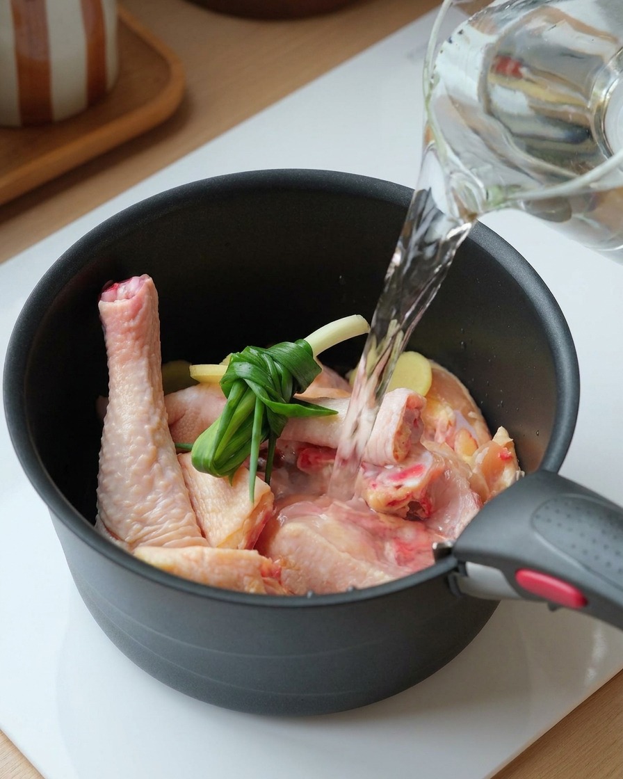 Clear water being poured from a glass pitcher into a black pot containing raw chicken pieces, tied scallions, and sliced ginger.