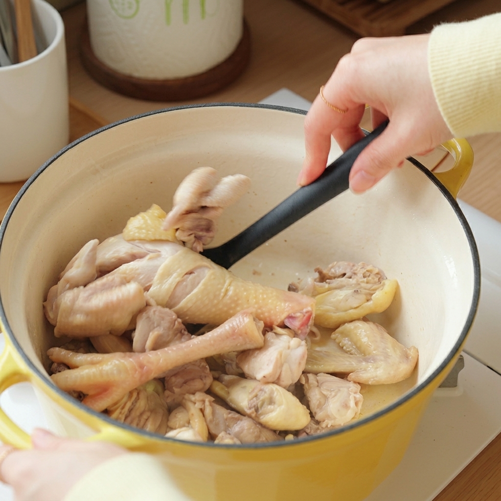 A hand using a black spatula to stir-fry blanched chicken pieces inside a yellow cast iron pot.