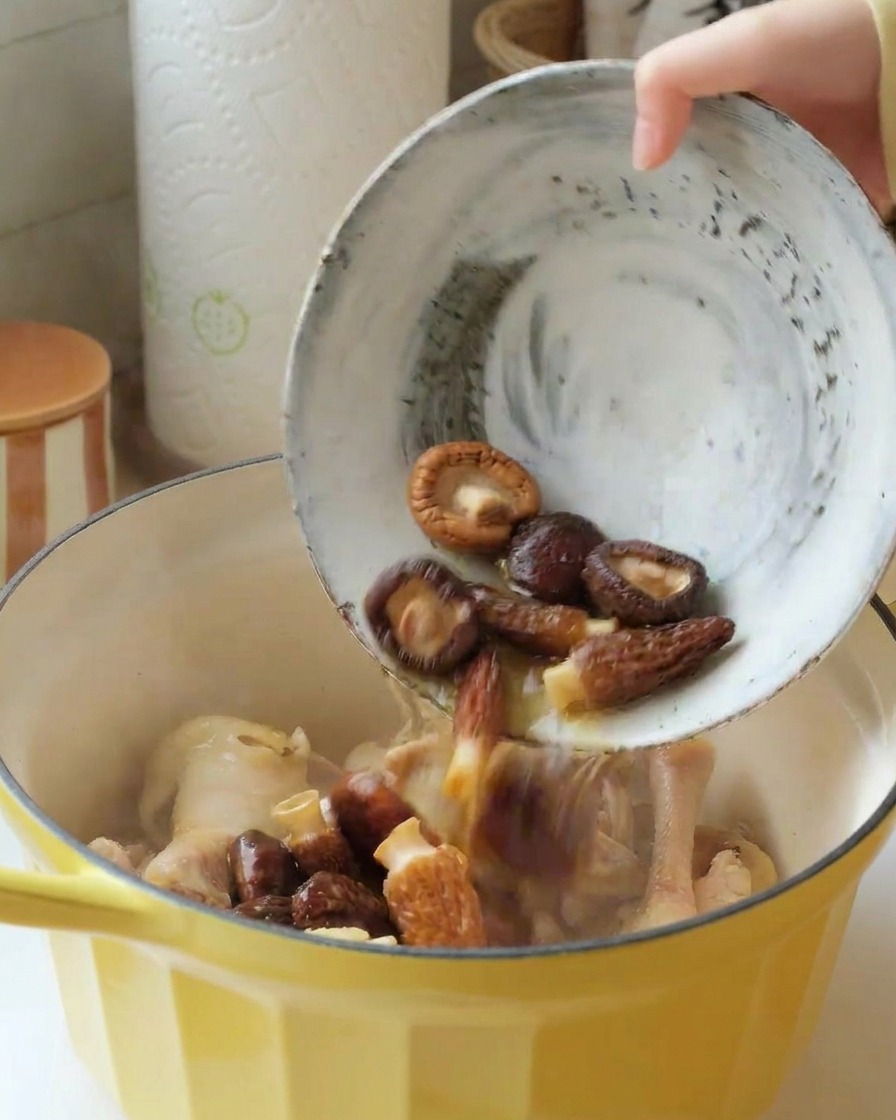 A bowl of soaked shiitake and morel mushrooms being poured into a yellow pot containing chicken pieces.