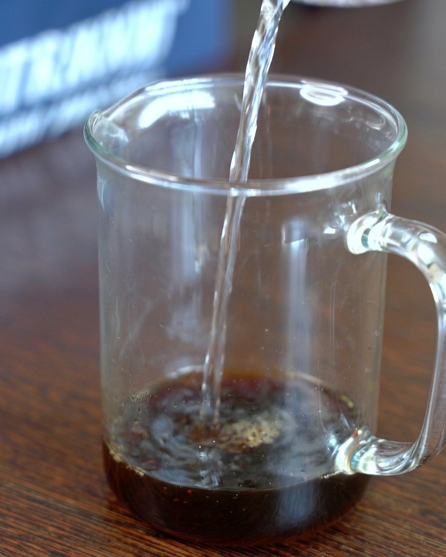 Clear water being poured into a glass measuring cup containing a dark mixture of kombu-infused soy sauce and sugar.