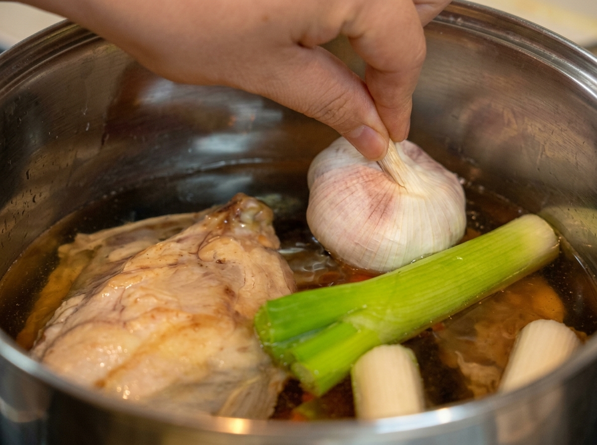 A hand placing a whole, unpeeled head of garlic into a pot filled with water, a chicken carcass, and green leek stalks.