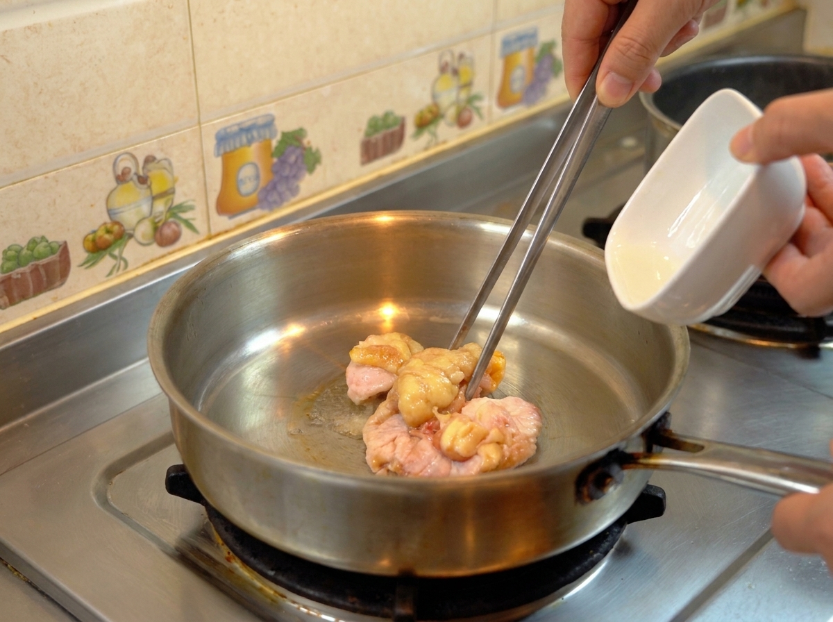Pieces of raw chicken fat being moved with metal tongs in a stainless steel skillet as they render into clear oil.