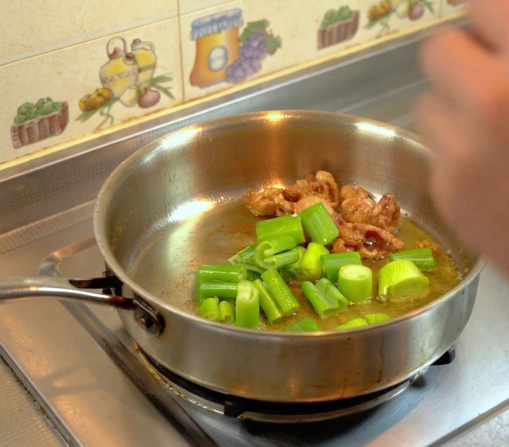 Thickly chopped green leek leaves frying in rendered golden chicken fat inside a hot stainless steel skillet.