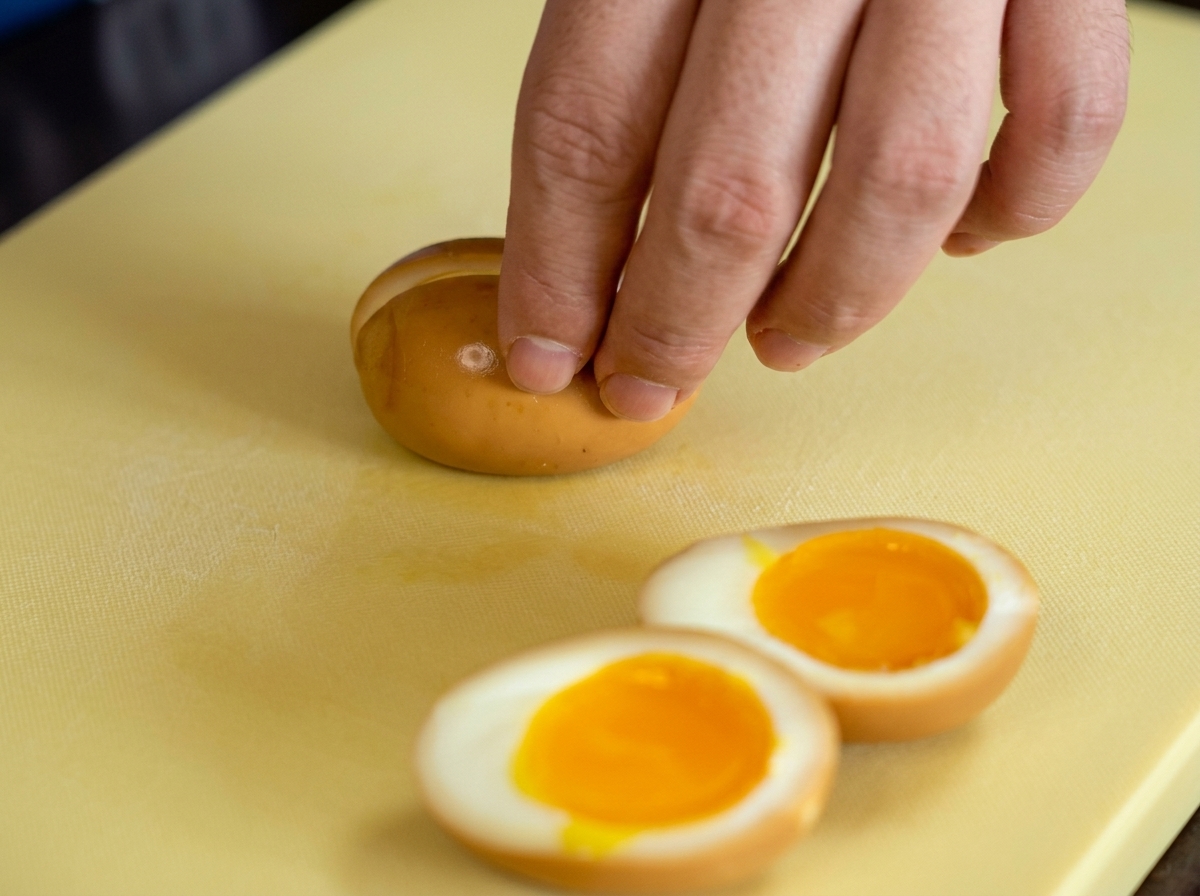 A hand slicing a brown, soy-marinated soft-boiled egg in half on a yellow cutting board, revealing a gooey orange center.